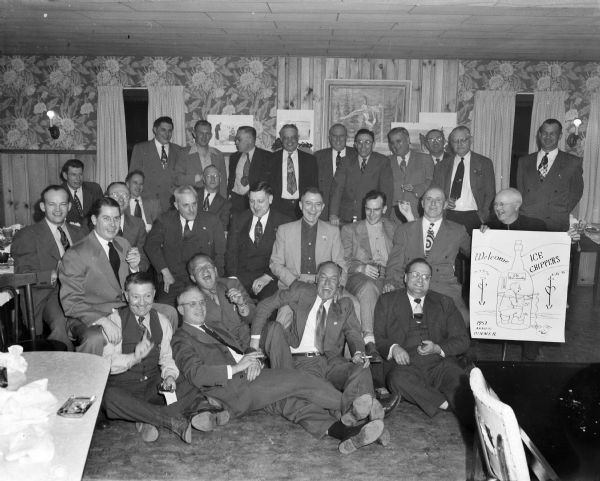 Group portrait of the "Ice Chippers," an ice fishing club of Gisholt workers gathered for their annual dinner. A man on the right is holding a hand-drawn poster of a man ice fishing from inside an Old Grandaddy whiskey bottle. Taped to the wall in the background are other comical posters created by Sid of men ice fishing.