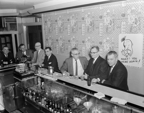Elevated view of men gathered at a bar before an Ice Chippers annual dinner. A poster on the wall depicts a bird with its head going through the sand so it is looking at its rear end with the message: "What! You here again!"