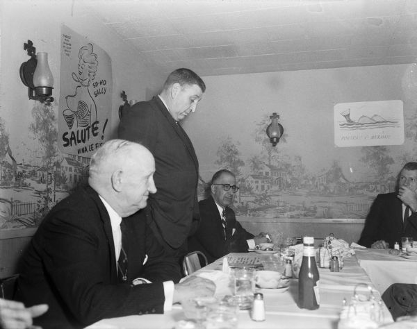 View down table towards a man standing and speaking to the group at an Ice Chippers annual dinner. Behind the men, posters are taped to the wall and include "Portrait of a Mermaid" and "So-Ho Sally." 