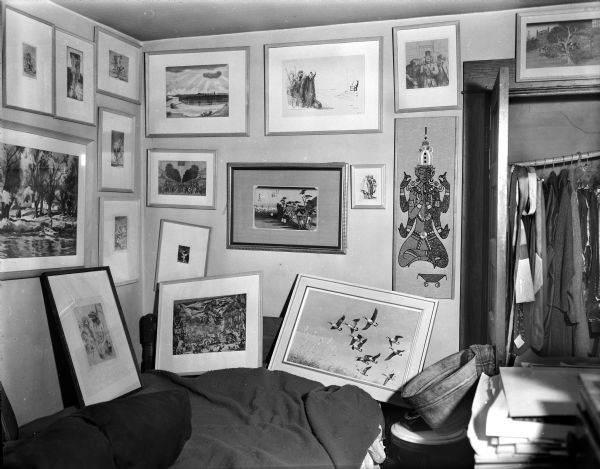 View of interior of Sid's bedroom with a number of paintings and other artworks framed and displayed on a wall in the corner of a room. There are clothes hanging in an open closet on the right. Some of the framed artworks are resting on a bed.