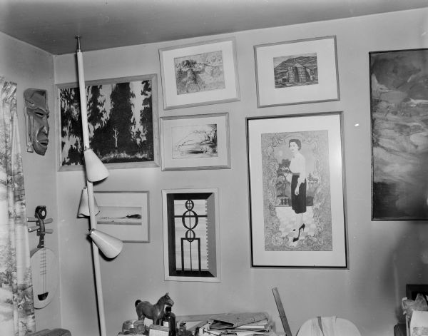 View of a corner of Sid's office with several artworks, including paintings, masks, musical instruments, etc. Near the corner is a floor to ceiling light fixture, and a table or desk piles with items.