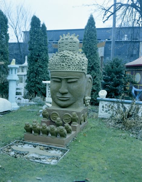 View of the sculpture, titled "Monumental Oriental Head" of a Buddha in the backyard of Sid's house. In the background in the yard are other sculptures, and the pagoda. In the far background is the Madison-Kipp Corporation factory. The dimensions of the painted cast concrete piece are 110" x 45" x 78".
