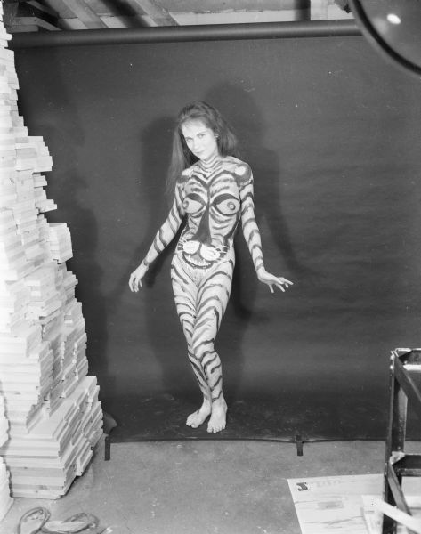 Full-length, black-and-white portrait of "Tiger Woman," a female model posing nude against a black paper backdrop. She is painted like a tiger with striped arms and legs, and the face of a tiger on her torso. Beside her on the left is a tall stack of cut wooden boards.