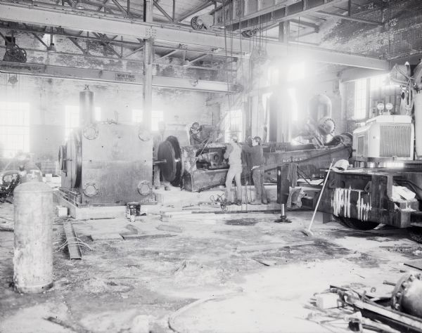 Group of men operating a machine suspended on chains attached to a crane inside the Gisholt factory.