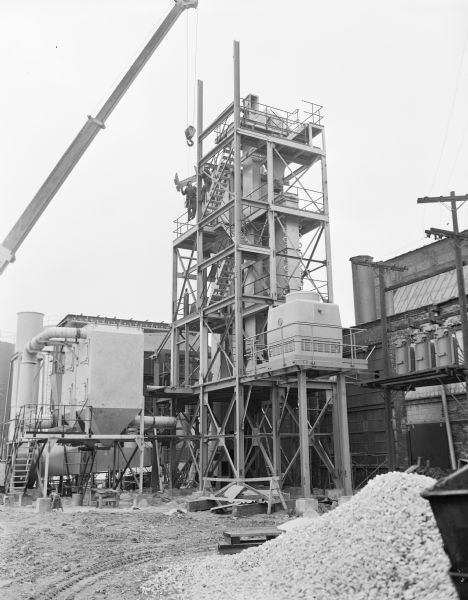 View outdoors across yard towards two workers standing near the upper level of an industrial tower. The men are guiding a structural part in place on the tower. There is a pile of gravel in the foreground, and in the background are factory buildings.