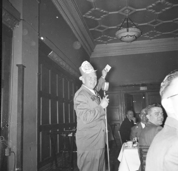 Sid Boyum wearing a hat with a sign for Gisholt attached to the front. Sid is standing and speaking at a Gisholt management club meeting. Men are sitting at tables on the right.