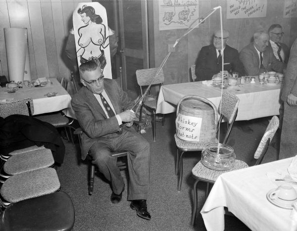 Elevated view of a man sitting in a chair fishing in a bowl of water at an "Ice Chippers" gathering. Next to him is a basket labeled: "Whiskey, Worms, What Nots." Behind him a man is posing behind a hand-drawn cutout of a topless woman. Other men sit at tables in the background. Comical drawings created by Sid hang on the wall.