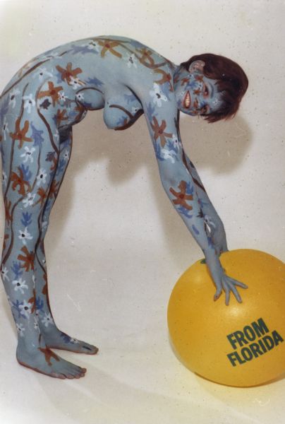 Side view of "Beach Ball Girl" posing in front of a white backdrop. She is wearing a base of blue body paint, with red, blue and white flowers, and abstract dark red lines on top. She is leaning over and resting her hands on an inflatable orange ball with "From Florida" printed on it.