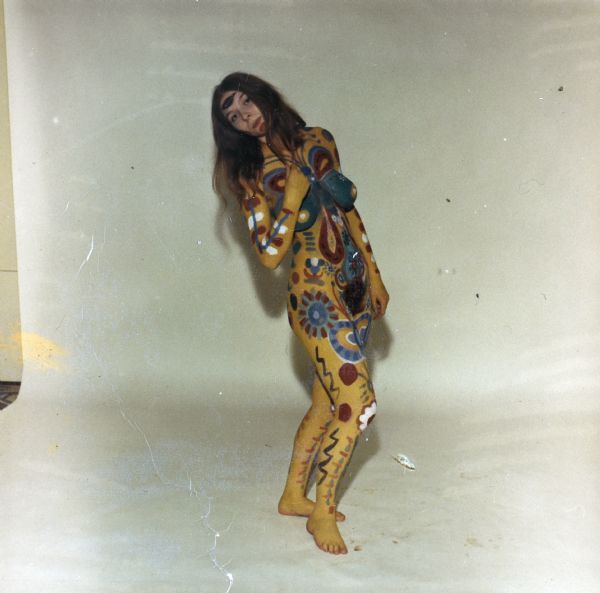 Full-length portrait of a nude woman wearing body paint of abstract and floral designs, posed standing in front of a white backdrop.