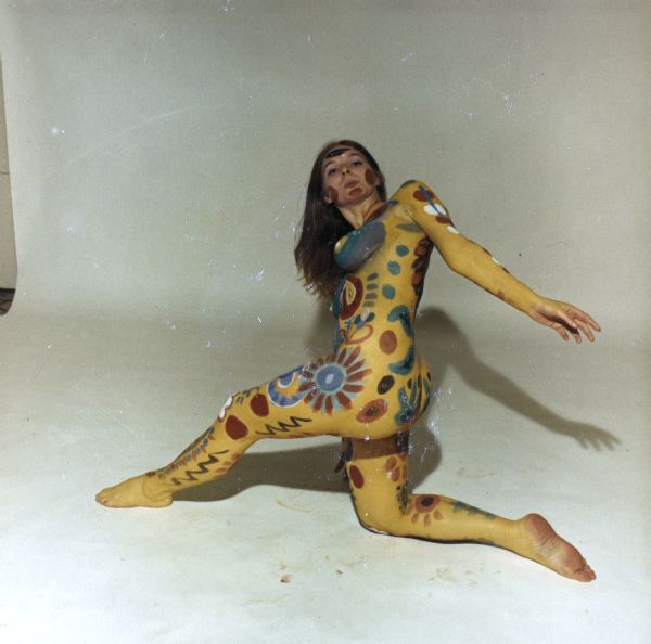 Full-length portrait of a nude woman wearing body paint of abstract and floral designs. She is posed in front of white backdrop, kneeling on her right knee, with her left leg stretched out to the left, her left arm stretched out to the right, and her head and torso turned toward the front.