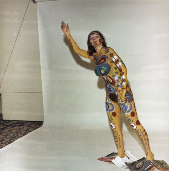 Full-length portrait of a nude woman wearing body paint of abstract and floral designs. She is posed standing in front of a white backdrop, and has magazine covers over her ankles.
