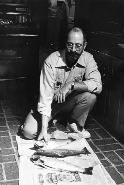 Steve Hopkins kneeling on a kitchen floor, looking at three gutted trout laid out on newspapers. He is wearing a dress shirt, khakis, white tennis shoes and a wristwatch.