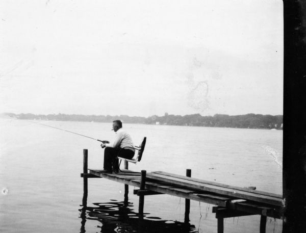 View across water towards Sid, with a cigar in his mouth, sitting on a chair on a pier and fishing in a lake. The far shoreline is in the background.