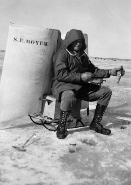 A man wearing boots and coat is sitting on a box on a sled. Behind him is a cloth (perhaps a wind break) with "S.E. BOYUM" printed on it. The man is holding a fish in his left hand.