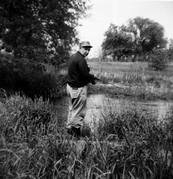 View of Sid fishing in tall grass at a pond or river. He is looking over his shoulder towards the camera.