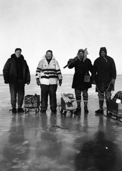 Group portrait of Sid Boyum and three other men, likely of the Gisholt Ice Chippers fishing club, standing on the ice with sleds and fishing equipment. 
