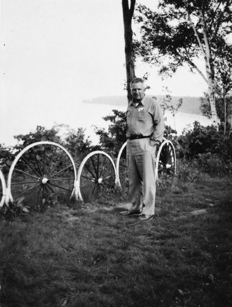 Portrait of Sid standing on a lawn near a row of wagon wheels set into the ground. A lake is below in the background. Sid is wearing khaki pants, and a button-up shirt, and has photographs in his shirt pocket.
