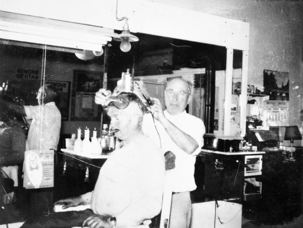 Sid is sitting in a barber's chair with a cigar in his mouth, and the barber standing behind him. The barber is holding some kind of hat contraption over Sid's head. There is a mirror with their reflections in it on the wall in the background. A calendar on the back wall on the right is from Meyers Drugs.