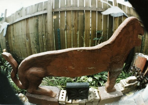 Fisheye lens view of an elongated lion sculpture in Sid's backyard. Behind the sculpture is a wire fence, and a taller wood fence, on which plywood bird cutouts have been placed.