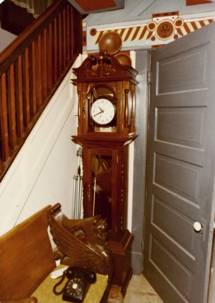 Sid Boyum's entry hall, with grandfather clock, carved wooden dragon head bench, Egyptian motif painted paneling, and old black rotary dial telephone. There is a door to kitchen on the right, and on the left is the railing for a staircase to the second floor.