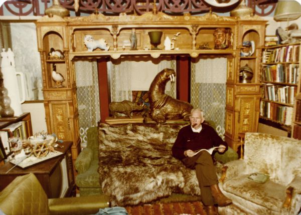 Sid Boyum, with a cigar in his mouth, is sitting on a fur-covered sofa in his living room reading a magazine. There is a large, carved wooden shelving unit behind him framing a bay window. There is a bookcase on the left, and rhinoceros and walrus sculptures are on the shelf above Sid's head.