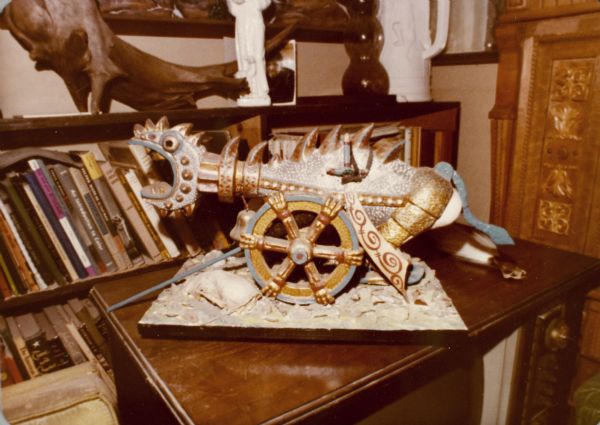 Wooden scultpure of a dragon-styled cannon, painted and decorated fancifully, resting on wood cabinet-style television in Sid's living room. There is a bookcase in the background.