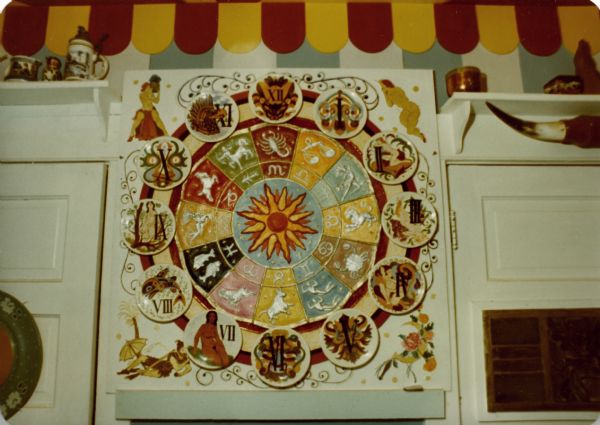 Non-functioning clock on the wall of Sid's kitchen. The clock consists 12 ceramic plates denoting each hour in Romain numerals and decorated with neoclassical figures, Germanic folklore and Haida-like fish. The plates form a clock face encircling an inner bas-relief of the sun. Radiating from the sun to the numerals are two concentric circles with brightly painted symbols of the Zodiac. The entire piece is mounted on a square plywood board with Greco-Roman figures painted in three of the four corners, the fourth having a floral design.