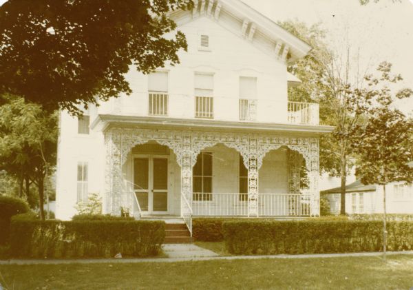 View from street of the front of a white, two-story house with pitched roof and front porch with decorative ironwork surround painted white. The house (in New Orleans?) has a second-story porch wrapping around the front and one side. There is a low, trimmed hedge in front along the sidewalk.