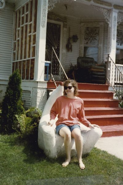 Outdoor portrait of a young woman sitting on the "Polar Bear Chair," a concrete cast sculpture in Sid's front yard. She is wearing sunglasses, shorts and sandals. Behind her is the front porch of Sid's house, with decorative "stained glass" panels, and fancy ironwork scrolls.