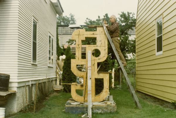 Sid Boyum, with a cigar in his mouth, is standing on a ladder while building a wooden form for his concrete casting of an abstract geometric sculpture that stands 118" x 62" x 58". The form is in a side yard between Sid's house and his neighbor's. The Madison-Kipp Corporation factory building is in the background.