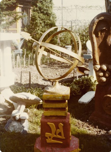 Metal astrolabe mounted on concrete base in Sid's backyard. The base is painted red with yellow Chinese characters, and the astrolabe is painted gold, with an arrow bisecting the globe. A garden bed and other sculptures are in the background.