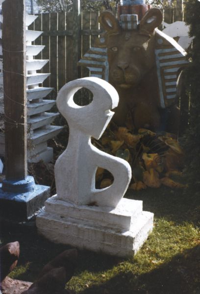 Sun-lit sculpture, identified as "Creature Abstract," in Sid's backyard. Painted entirely white, the sculpture uses bold negative spaces with curvilinear shapes to define an abstract human figure: a small head with ocular and a nasal or mouth cutout sits on an upper torso, defined by shoulders and open arm or breast. The body stands on a two-tiered base, and in its entirety measures 47" x 31" x 20". Other sculptures pictured are "Sphinx," a white "Multi-tiered Pagoda," and a pillar.