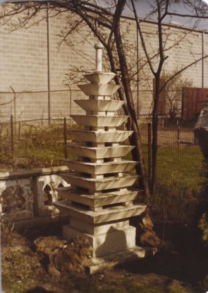 White-washed, cast-concrete sculpture identified as "Multi-tiered Pagoda" and measuring 92" x 32" x 32". It stands in Sid's backyard in front of a tree. Two sections of low white border fence is behind the sculpture. The brick wall of Madison-Kipp Corporation is in the far background. 

A handwritten notation of Sid's on the back of an undigitized image states that the pagoda took two-and-a-half tons of cement to make. 

