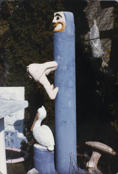 Tall blue pillar identified as "Odd Tower" with a grinning face at the top. A figure partway down has its head inside the pillar and feet braced against the pillar. At the base is a white pelican sitting on a blue base. To the right at the base of the pillar is the small sculpture known as "Smiling Mushroom."