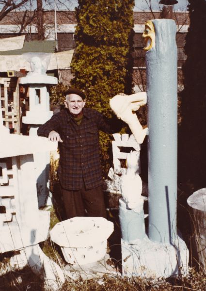 Sid Boyum standing in his backyard between the Blue Pillar with Figure and Pelican, and a large white Lantern sculpture. Other sculptures are in the background. In the far background is Madison Kipp Corporation.