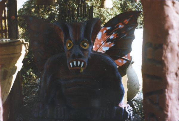 High-relief, concrete-cast sculpture of a winged, four-armed gargoyle in Sid's backyard. The mouth is gaping with white fangs. Its upper body and head are painted purple and red.