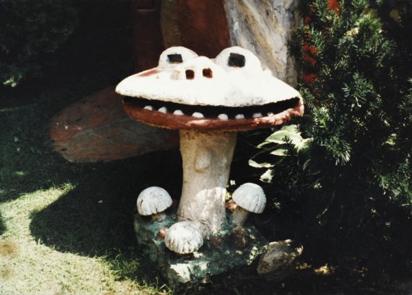 Short cast-concrete sculpture identified as "Smiling Mushroom." It is an anthropomorphized form having a yellow face with eyes and nose on top of the mushroom cap; a red mouth with white teeth opens on the side of the cap. The green base is square and has three small white mushrooms. In the background is the base of another sculpture, shrubbery and a hosta plant. It measures 26" x 24" x 32".