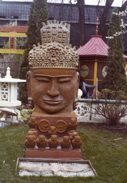 Large concrete-cast sculpture painted red and titled "Monumental Oriental Head" of a Cambodian-styled Buddha with heavily textured light-colored hair and topknot. With dimensions of 110 x 45 x 78 inches, it sits on a thick base ornamented with round Zodiac symbols in Sid's backyard. A group of molded owls surround the space in front. On the ground before the sculpture is a flat goose shape, wings outstretched, on a bed of gravel. Part of a white lantern and torii gate appear are on the left, and the tea house is in the background on the right. A Madison-Kipp Corporation building is in the far background.