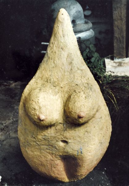 Semi-abstract, yellow cast concrete sculpture, called "Female Teardrop." Similar to prehistoric "Venus" figures, it has prominent breasts and wide hips. The top comes to a point instead of a head. 