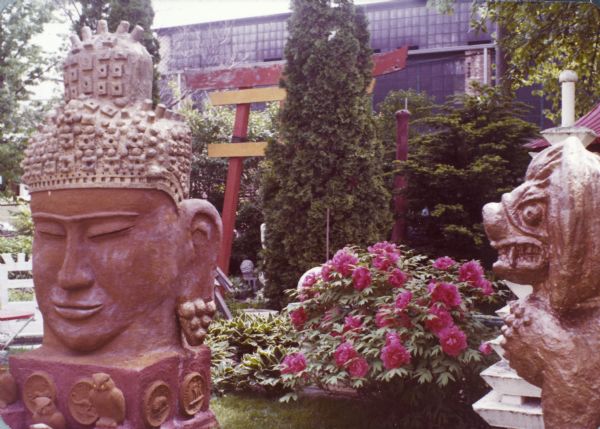 Sid's backyard with Cambodian-styled sculptures titled "Monumental Oriental Head" of a Buddha and "Stylized Oriental Lion." The head stands 110" x 45" x 78" and the lion, 61" x 35" x 28". In the center is a large flowering peony bush, hosta, cedar trees, and torii gate. Madison-Kipp Corporation is in the background.