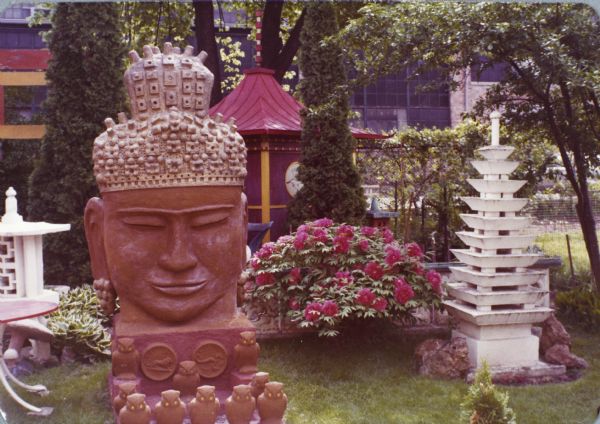 Sid's backyard with Asian-themed, concrete-cast sculptures titled "Monumental Oriental Head" of a Buddha (with dimensions of 110" x 45" x 78"), "Multi-Tiered Pagoda" (92" x 32" x 32" to the right), white lantern (to the left), portion of the torii gate (in far right background), and the pagoda with a red sloped roof. The Buddha head, inspired by early Cambodian styles, sits on a thick base ornamented with round Zodiac symbols. A group of molded owls surround the space in front.In the center is a large flowering peony bush, hosta, cedar trees, and torii gate. Madison-Kipp Corporation is in the background.