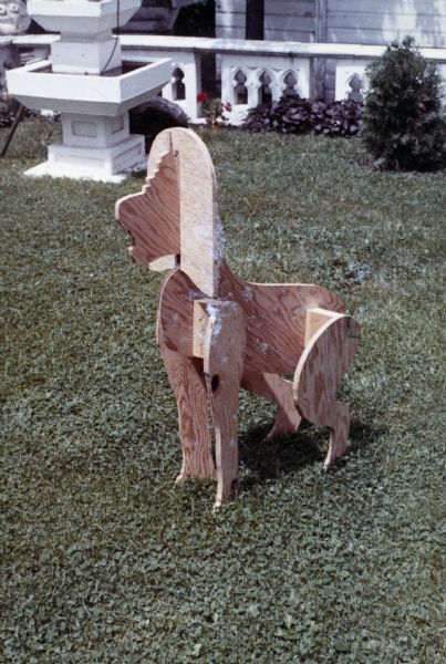 Armature made of flat, interlocking plywood pieces sitting on the lawn in Sid's backyard, in the general shape of the Cambodian Lion sculpture. Parts of the White Fountain and a church railing are in the background in front of a building.