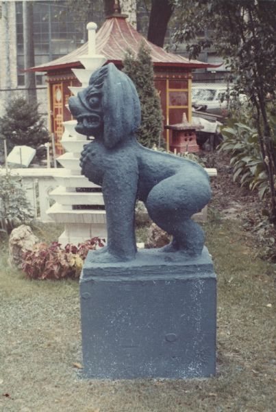 Southeast Asian-styled "Guardian Lion" concrete-cast sculpture on a large rectangular base painted blue in Sid's backyard. In the background is the white "Multi-tiered Pagoda" and the tea house. Madison-Kipp Corporation is in the background.