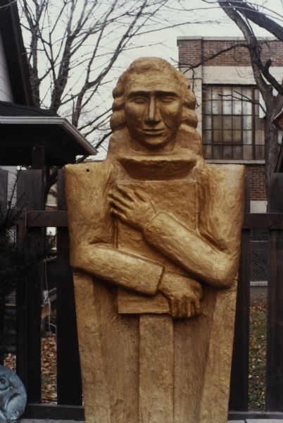 High relief sculpture of a man in an academic robe clutching a book to his chest, standing against a fence. In the background is the house next door and Madison-Kipp Corporation.