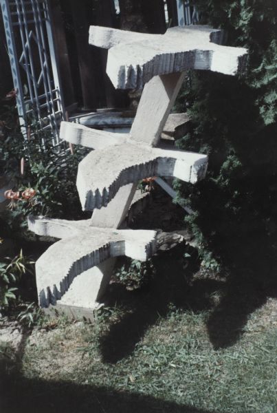 Three flat goose shapes, with wings outstretched, one above the other on a square post set at an angle, set in the yard among garden plants, shrubs, and a white trellis leaning against a building.