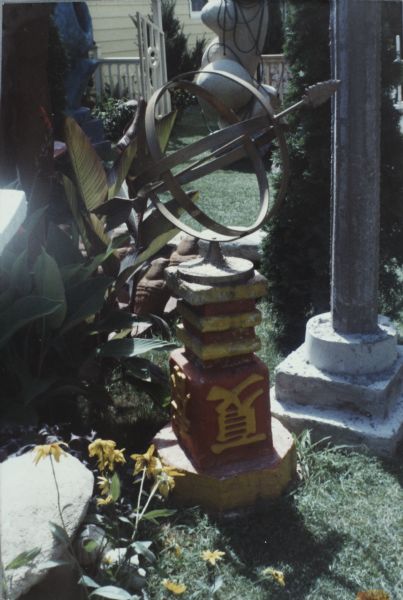 Metal astrolabe mounted on concrete base in Sid's backyard. The base is painted red with yellow Chinese characters, the  Astrolabe is painted gold. In the background are sculptures, plants, shrubs, lawn, and a building behind a fence.