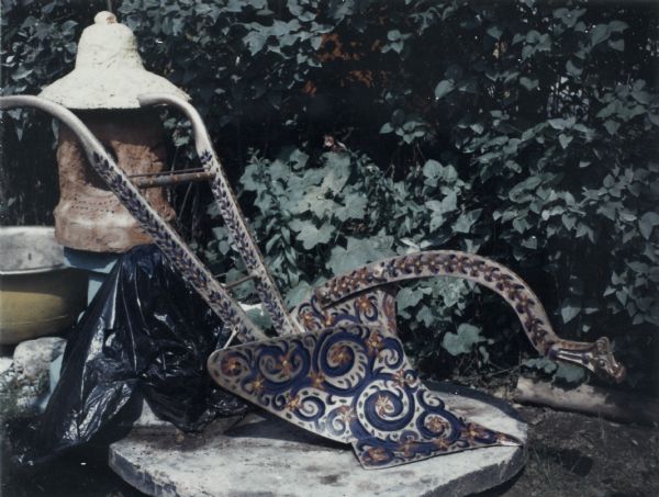 A plow with handles, plowshare and hitch is painted with multicolored design of spirals, dots and chevrons. A red concrete lantern is on the left, and leafy vegetation is in the background.