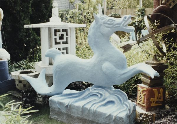 White horse sculpture with fish tail on a base of swirling water forms, in front of the Astrolabe and a medium-sized white lantern in Sid's backyard.