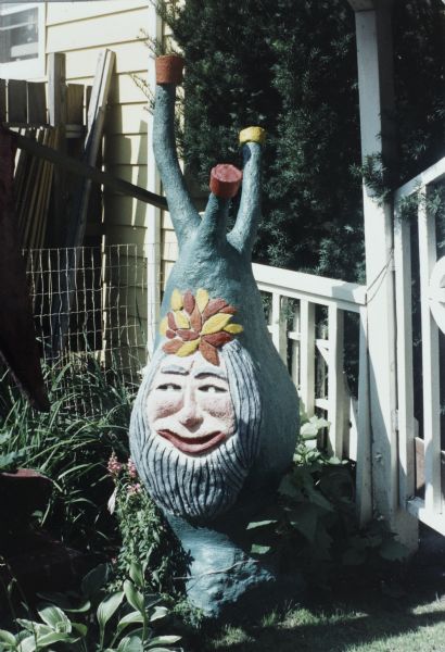 Blue-green, pear-shaped sculpture on a round base with three limb-shapes on top in  Sid Boyum's backyard. The face is painted in natural colors with incised lines for hair and beard. Forehead topped with yellow and red flowers. Limb shapes topped with yellow and red. Surrounded by garden foliage and a white fence, and a building in the background.