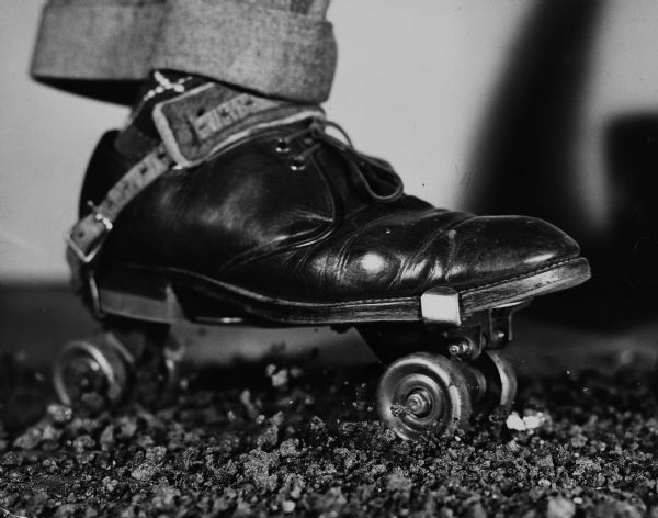 Illustrative art showing a close-up of a man's dress shoe strapped onto a roller skate with leather bindings. A pants cuff and hint of an argyle sock are visible at the ankle. A few rocks are airborne under the skate and by the front wheel to show motion. 